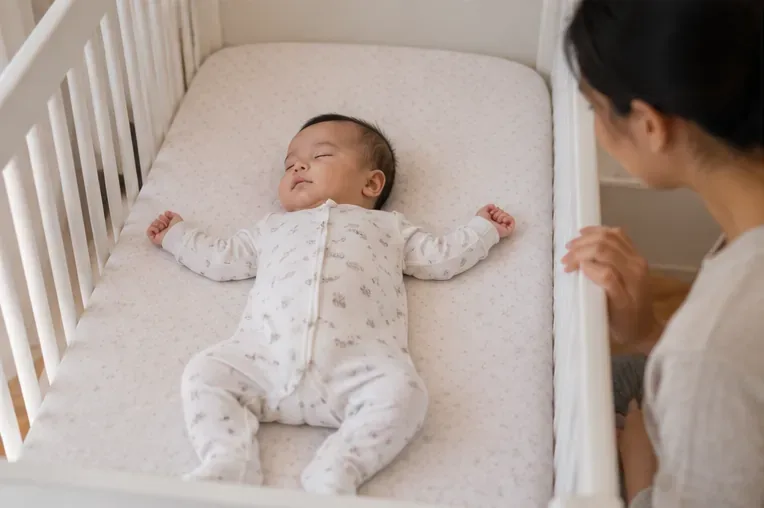 An infant sleeps on their back in a bare crib on a flat mattress while a parent checks nearby.